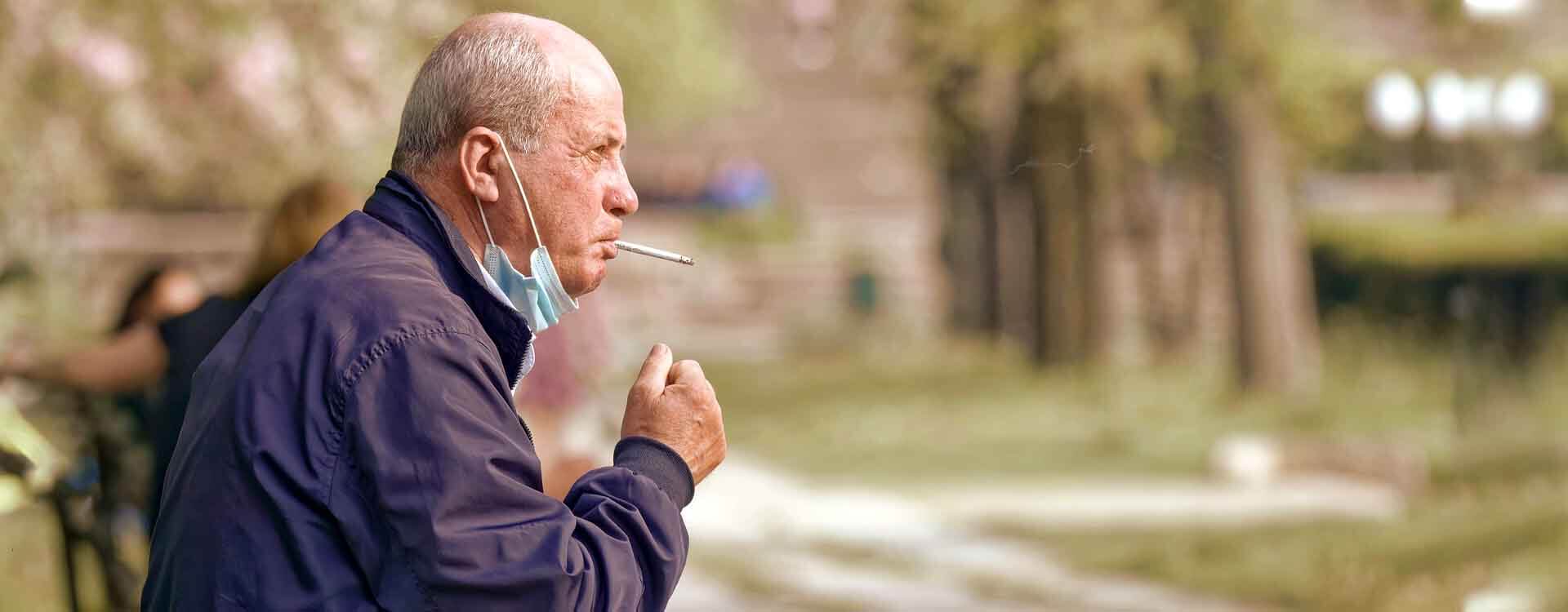 Starší pán fajčí cigaretu v parku so zloženým rúškom