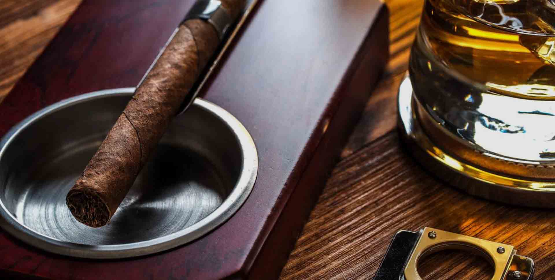 Cigarový popolník - detail: cigara odložená v popolníku, časť orezávača a pohárika s alkoholom