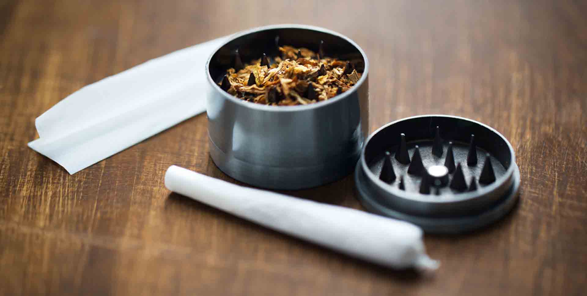 Kovový grinder plný tabaku, papierik na cigarety a ubalená cigareta